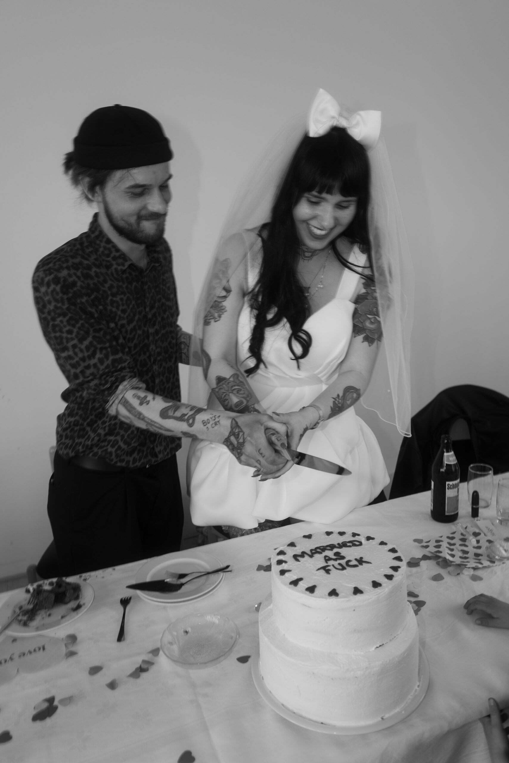 Braut & Bräutigam schneiden gemeinsam eine Hochzeitstorte an, lachen, tätowierte Hände, unkonventionelle Hochzeitsfotografie.