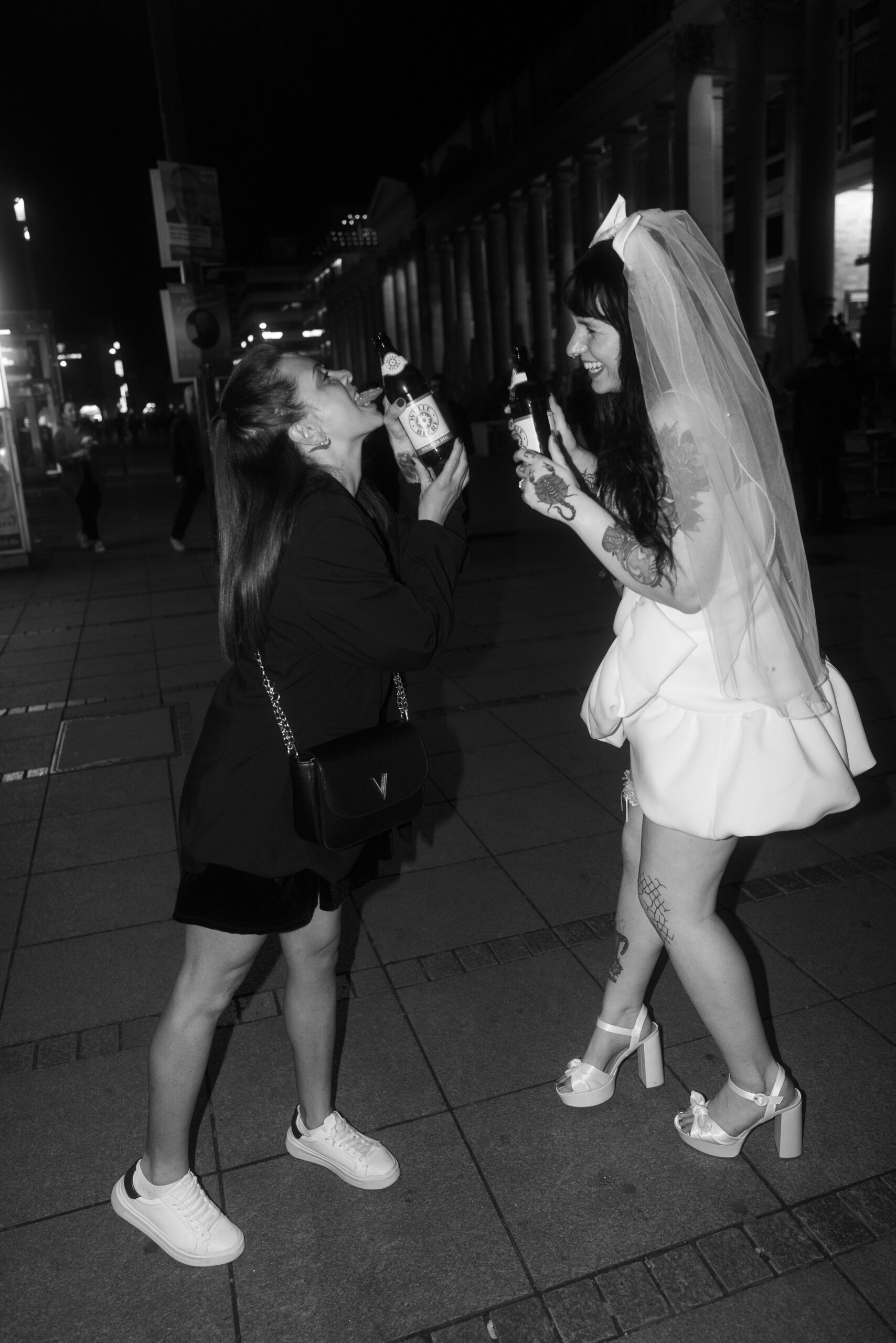 Braut & Freundin trinken zusammen auf der Straße, alternative Hochzeitsreportage, echte Momente.