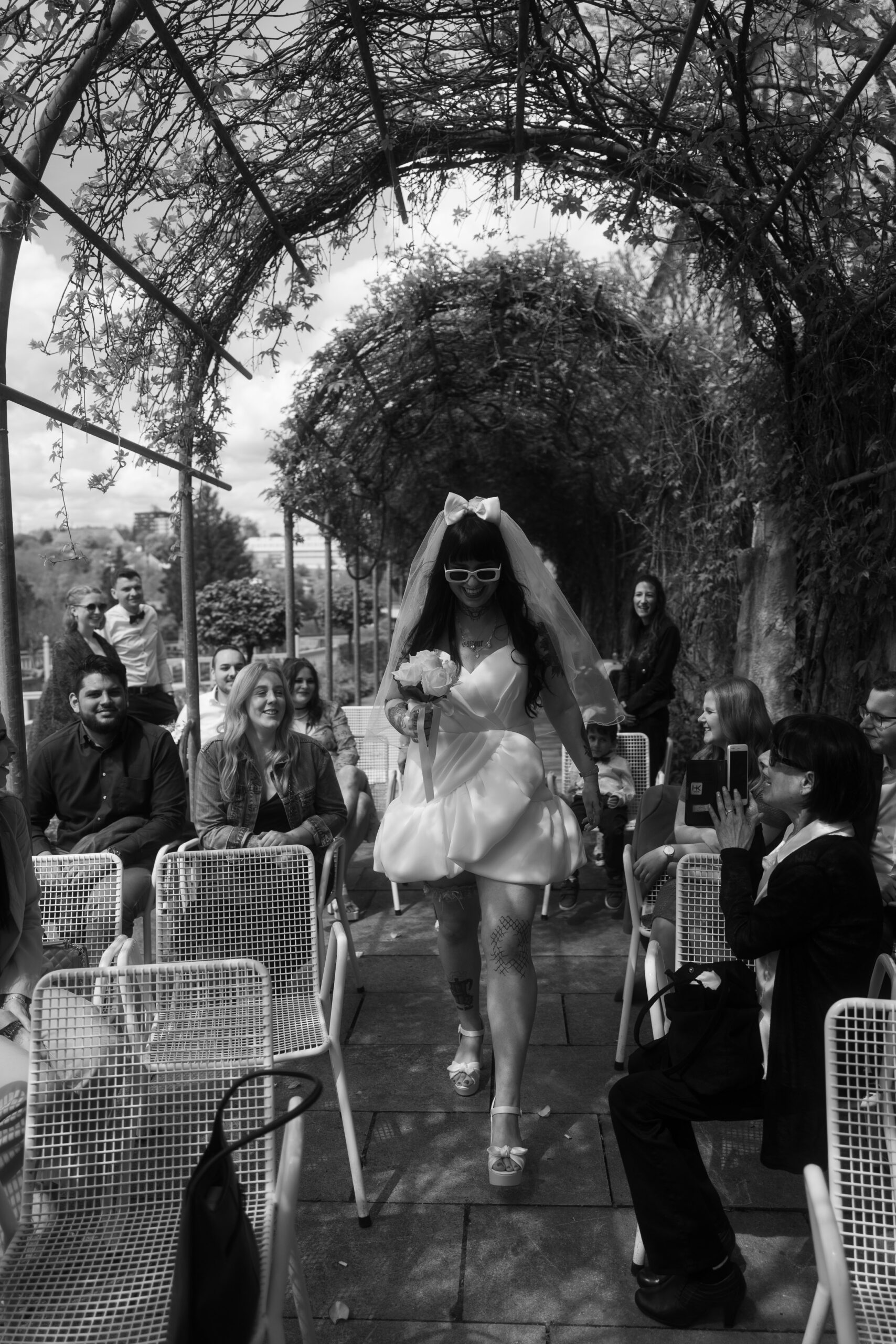 Braut mit kurzem Hochzeitskleid läuft voller Energie durch den Hochzeitsgang, Gäste lachen – unkonventionelle Hochzeitsfotografie.