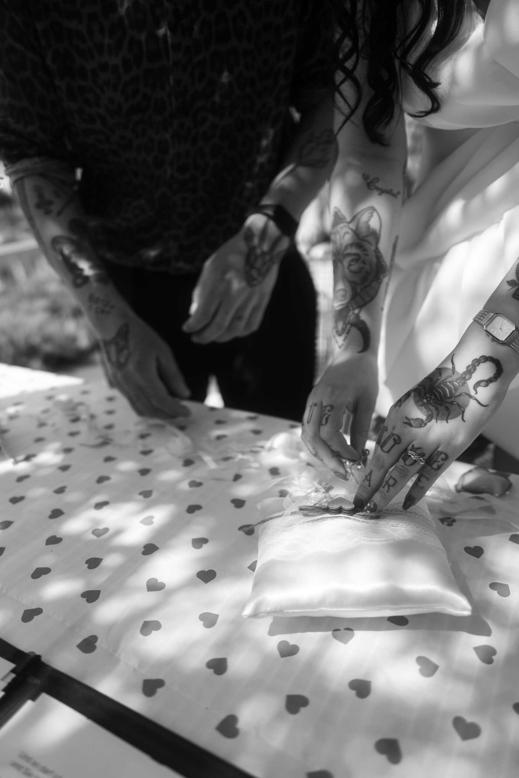 Tätowierte Hände von Braut & Bräutigam greifen nach den Eheringen auf einem Ringkissen, intime Momentaufnahme.