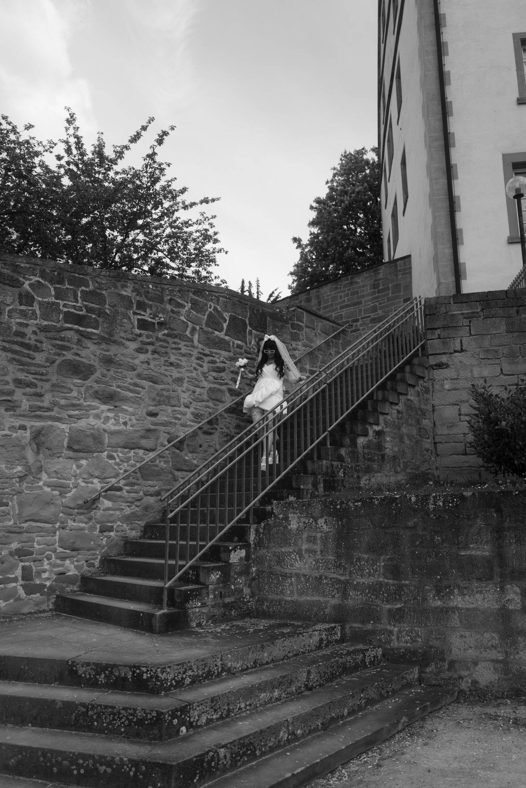 Braut in Sonnenbrille und kurzem Hochzeitskleid mit Blumenstrauß geht eine historische Steintreppe hinunter, umgeben von alten Mauern und Bäumen.