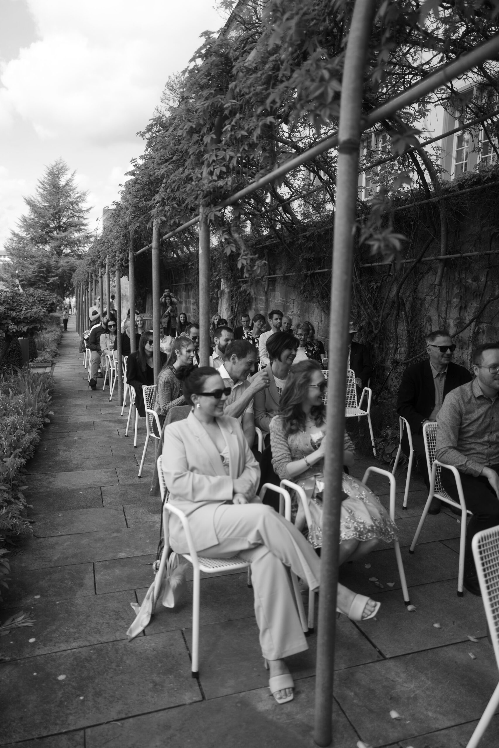 Hochzeitsgäste in Sonnenbrillen sitzen entspannt unter einem Laubengang – unkonventionelle, entspannte Hochzeitsfotografie.