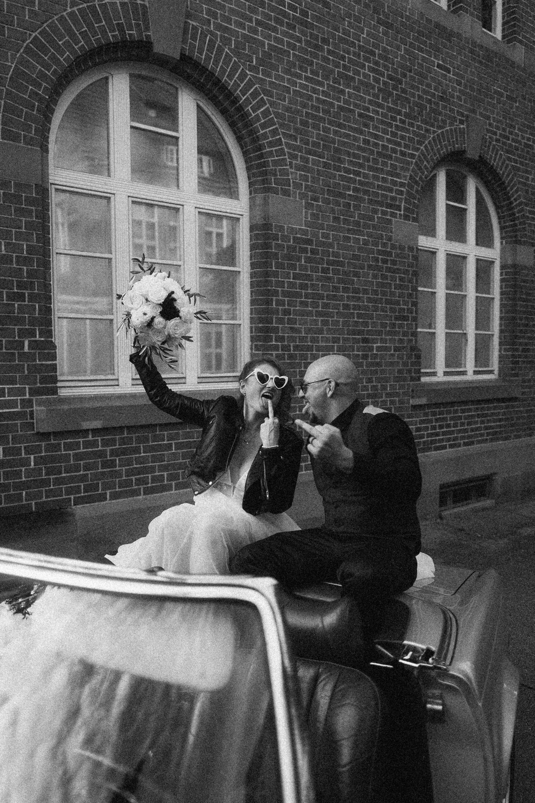 Braut mit Herzsonnenbrille und Mittelfinger-Pose auf Oldtimer mit Bräutigam – rebellisches Brautpaar bei Hochzeit im Da Capo Stuttgart