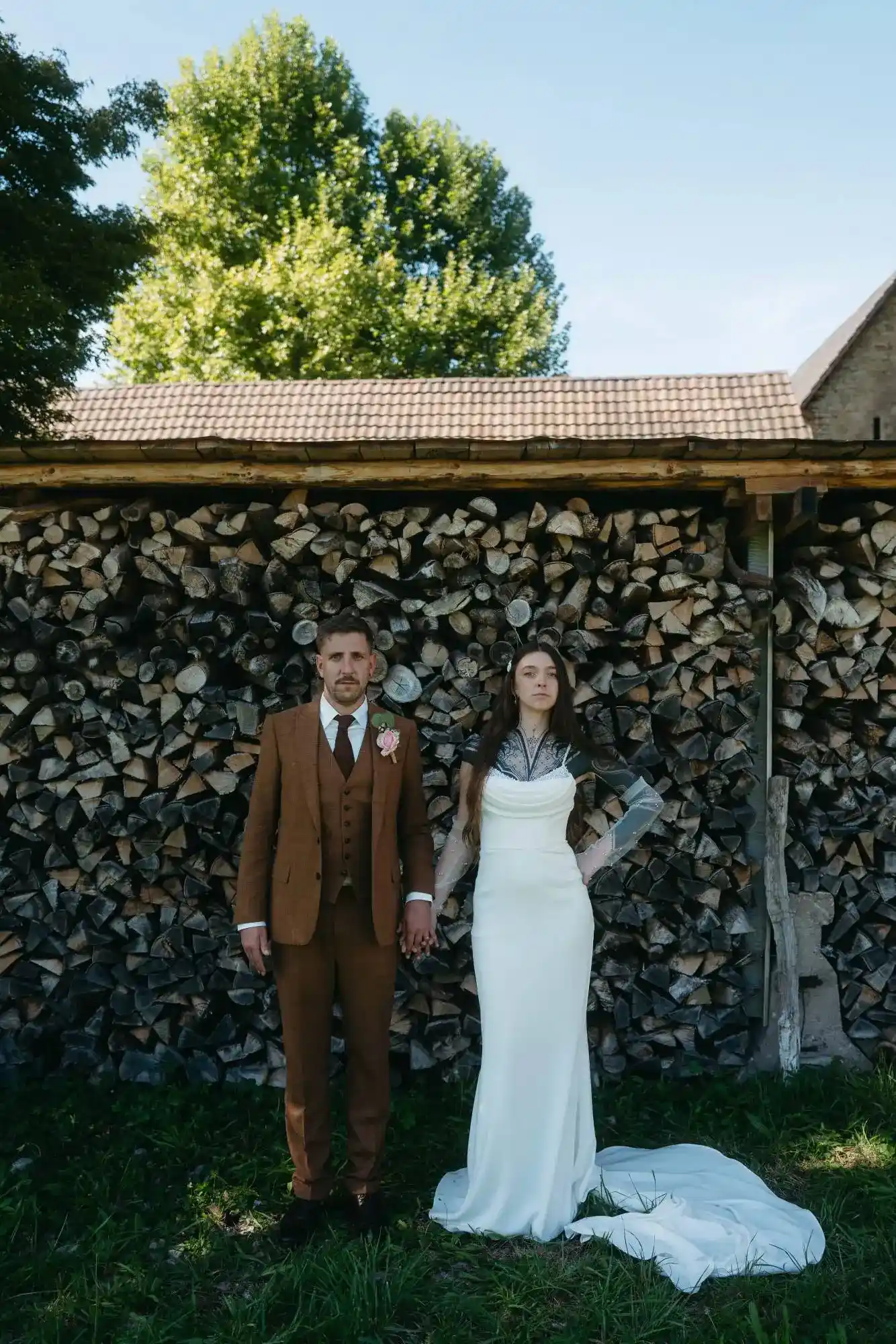 Braut und Bräutigam posieren vor gestapeltem Brennholz – ein modernes Porträt mit rustikalem Charme und starker Haltung.