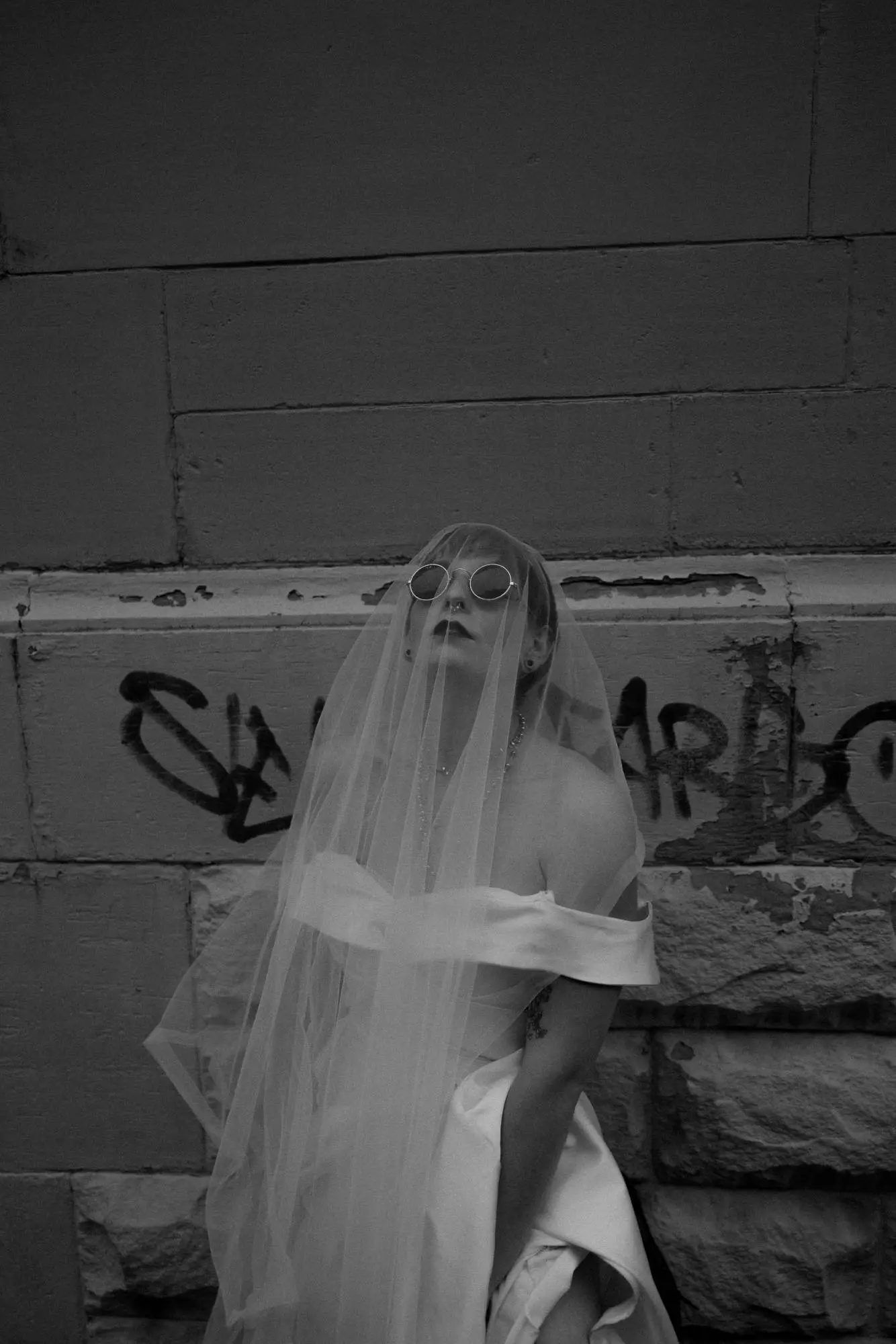 Braut in Sonnenbrille und Schleier steht vor einer Graffiti-Wand. Moderne dokumentarische Hochzeitsfotografie zwischen Eleganz und urbaner Attitüde.