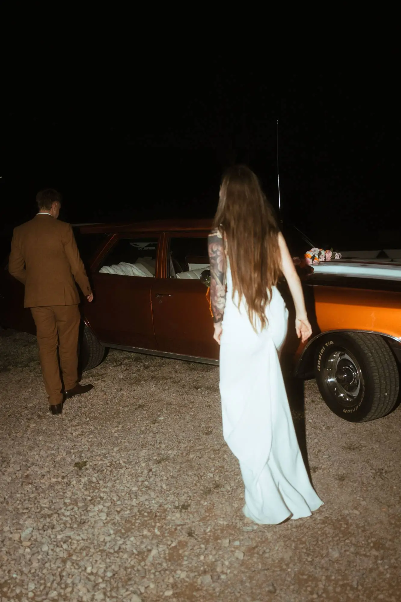 Braut in schlichtem Kleid geht bei Nacht gemeinsam mit dem Bräutigam auf einen braunen Vintage-Oldtimer zu.