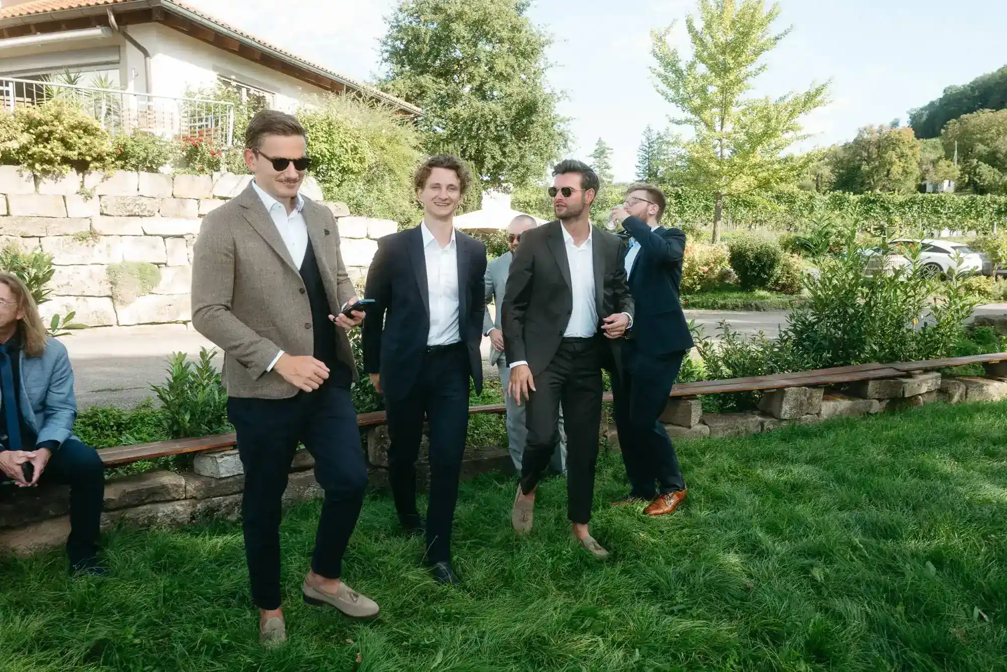 Vier stilvoll gekleidete Hochzeitsgäste im Anzug lachen gemeinsam in lockerer Atmosphäre im Grünen – moderne dokumentarische Hochzeitsfotografie fängt spontane Momente ein.