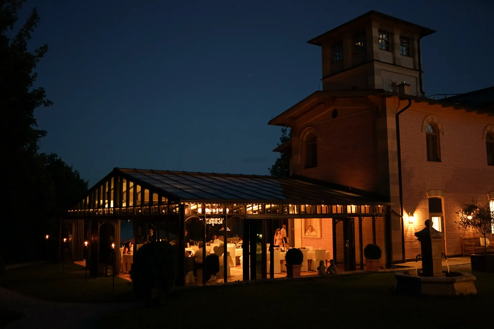 Stimmungsvoll beleuchtete Hochzeitslocation bei Nacht mit verglastem Wintergarten und warmem Licht, das nach draußen strahlt.